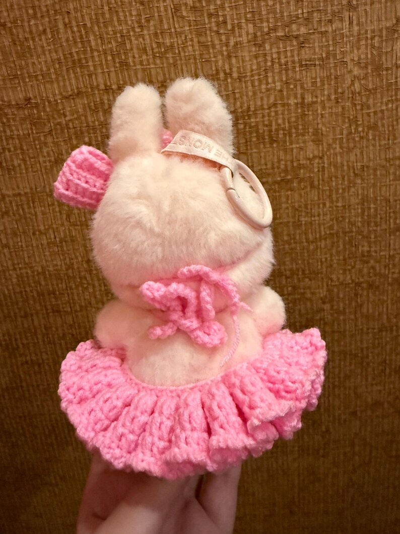 May include: A plush pink bunny toy with a crocheted pink tutu and bow. The bunny has a tag that reads "THE MONST". The toy is soft and fluffy, with a cute and whimsical design.