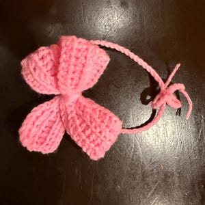 May include: A handmade, pink crocheted flower with a long, thin stem and a small, star-shaped detail at the end. The flower has a textured appearance, with visible stitch patterns. The item is likely a decorative element or embellishment.