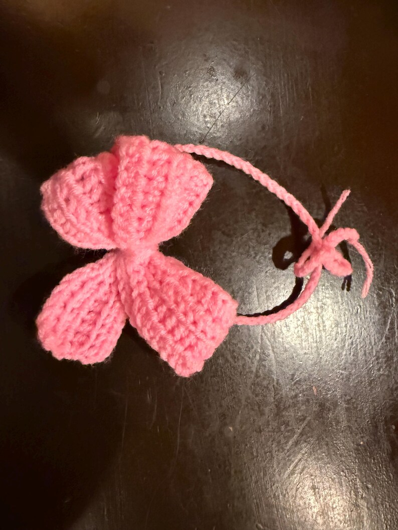 May include: A handmade, pink crocheted flower with a long, thin stem and a small, star-shaped detail at the end. The flower has a textured appearance, with visible stitch patterns. The item is likely a decorative element or embellishment.