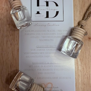 Könnte beinhalten: Drei transparente Auto-Lufterfrischer aus Glas mit Holzaufsätzen und Aufhängeschnüren werden neben einer weißen Karte mit schwarzem Text präsentiert. Auf der Karte steht "Fine Design Luxury Candles" und weiterer Text auf Deutsch.