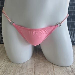 May include: A light pink thong bikini bottom with thin straps and silver metal rings. The bikini bottom is displayed on a white mannequin. The fabric appears smooth and slightly shiny.