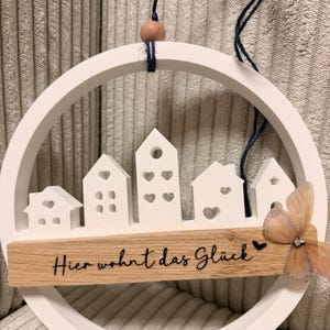 Könnte beinhalten: Weißer, kreisförmiger Wandbehang aus Holz mit einer Miniatur-Stadtszene. Er hat kleine weiße Häuser mit herzförmigen Ausschnitten, eine Holzbasis mit dem Text "Hier wohnt das Glück" und einen dekorativen Schmetterling.