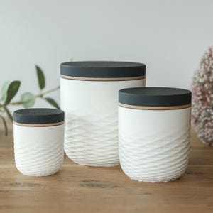 Op de afbeelding: Drie witte keramische potten met zwarte deksels en een textuur van golven. De potten zijn van verschillende formaten en staan op een houten oppervlak. Een bloemstuk en groen zijn op de achtergrond.