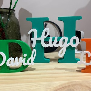 May include: Green, teal, and orange wooden letters spelling out the names "David", "Hugo", and "Lidia". The names are written in white cursive script. The letters are displayed on a wooden surface.