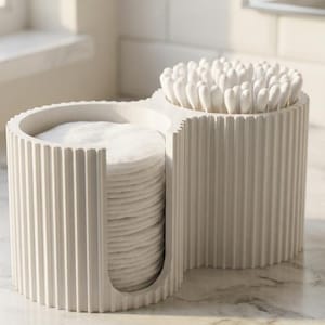 Può includere: Un contenitore bianco a due scomparti, scanalato, contenente dischetti di cotone e bastoncini. Il contenitore è realizzato in un materiale chiaro e poggia su una superficie di marmo. I dischetti di cotone sono impilati in uno scomparto.