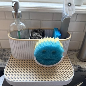 Op de afbeelding: Een gootsteenorganizer met een doorzichtige glazen zeepdispenser, een afwasborstel en een blauw-gele spons met een lachend gezicht. De organizer staat op een beige dienblad met een rasterpatroon.