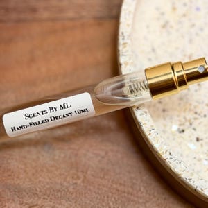 May include: A clear glass perfume decant bottle with a gold-colored atomizer. The label reads "Scents By ML Hand-Filled Decant 10ml". The bottle is on a wooden surface.