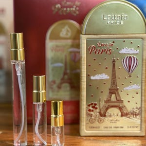 Pu&ograve; includere: Una collezione di flaconi di profumo e confezioni. I flaconi sono trasparenti con tappi color oro. La confezione comprende una scatola rossa e una scatola rettangolare color oro con la scritta "Love in Paris" e un'illustrazione della Torre Eiffel.