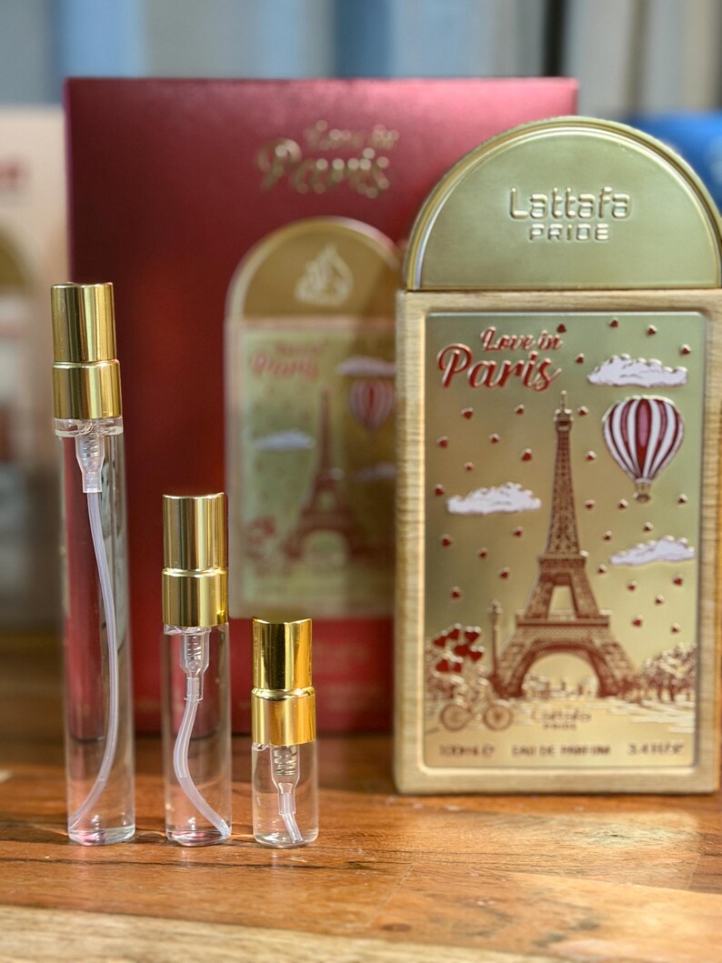 Pu&ograve; includere: Una collezione di flaconi di profumo e confezioni. I flaconi sono trasparenti con tappi color oro. La confezione comprende una scatola rossa e una scatola rettangolare color oro con la scritta "Love in Paris" e un'illustrazione della Torre Eiffel.