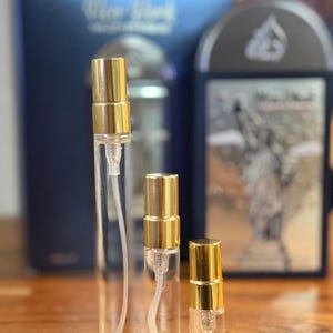 Pu&ograve; includere: Tre vaporizzatori di profumo in vetro trasparente con tappi color oro. I vaporizzatori sono di diverse dimensioni, con una scatola blu e un astuccio nero e argento sullo sfondo. I vaporizzatori sono su una superficie di legno.