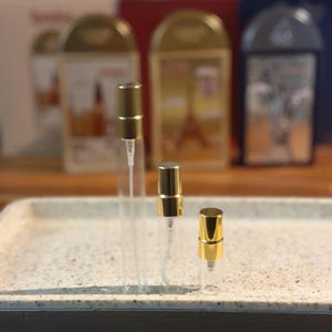 Pu&ograve; includere: Tre flaconi di campioni di profumo in vetro trasparente con vaporizzatori color oro sono disposti su un vassoio maculato. I flaconi variano di dimensioni. Sullo sfondo, ci sono tre scatole di profumo con dettagli dorati.
