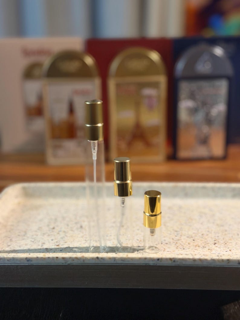 Pu&ograve; includere: Tre flaconi di campioni di profumo in vetro trasparente con vaporizzatori color oro sono disposti su un vassoio maculato. I flaconi variano di dimensioni. Sullo sfondo, ci sono tre scatole di profumo con dettagli dorati.