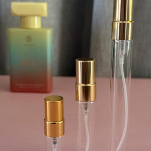 Puede incluir: Tres atomizadores de perfume de vidrio transparente con tapas y mecanismos de pulverización dorados. En el fondo, una botella de perfume rectangular más grande con una tapa amarilla y una etiqueta degradada. Las botellas están sobre una superficie rosa.