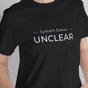 以下が含まれることがあります： 白い文字で「System Status - UNCLEAR」と書かれた黒いTシャツ。テキストは中央に配置されています。Tシャツは青いジーンズと合わせて着用されています。クルーネック。