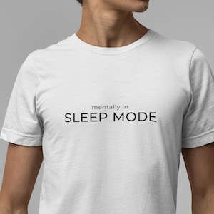 Könnte beinhalten: Weißes T-Shirt mit dem schwarzen Aufdruck "mentally in SLEEP MODE". Das Shirt hat einen Rundhalsausschnitt und wird mit einer blauen Jeans getragen. Das Gesamtthema ist Freizeitkleidung.