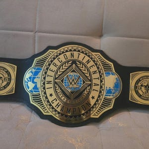 Puede incluir: Un cinturón negro y dorado del Campeonato Intercontinental. El cinturón presenta las palabras "Intercontinental Champion" y el logotipo de la WWE. Las placas laterales dicen "Never Give Up, Hustle, Loyalty, Respect, John Cena".