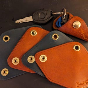 May include: Leather key holders in navy blue and brown with brass rivets. A car key and a leather key case are also present. The key holders are arranged on a dark surface.