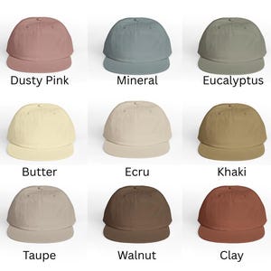 May include: Nine baseball caps in various colors, including dusty pink, mineral, eucalyptus, butter, ecru, khaki, taupe, walnut, and clay. Each cap has a curved brim and a rounded crown. The names of the colors are printed below each cap.