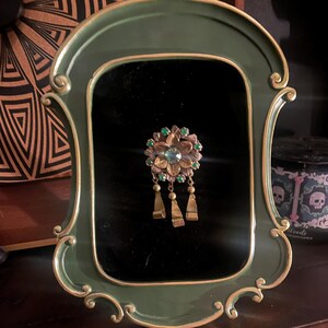 May include: An antique brooch with a floral design, featuring a central blue stone and green accents, hangs within a decorative green and gold frame. The brooch has dangling gold-toned pendants. The frame is set against a black background.