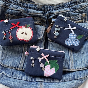以下が含まれることがあります： 紺色のコインケース3つ。それぞれに果物と花のアップリケが施されています。各ケースにはジッパー開閉と、チャーム付きの装飾ピンが付いています。1つはリンゴ、もう1つはブルーベリーのデザイン、3つ目はイチゴです。