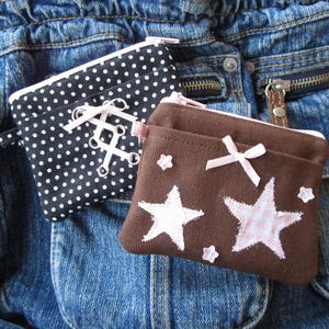 Peut inclure: Deux petites pochettes en tissu faites à la main. L'une est noire à pois blancs avec un détail de ruban rose. L'autre est marron avec des appliqués d'étoiles blanches et roses et un nœud rose. Les deux ont des fermetures éclair, sur un fond en jean.