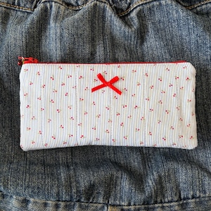 May include: A rectangular pencil case with a white background, blue vertical stripes, and small red cherry patterns. A red bow is centered on the case. The zipper is red.