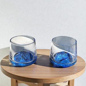 May include: Two clear glass tumblers with a blue mountain design at the base. The glasses sit on a round wooden table. The blue design is visible through the clear glass, creating a unique visual effect.