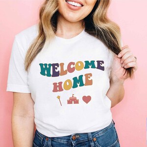 Könnte beinhalten: Weißes T-Shirt mit dem Text "WELCOME HOME" in bunter Retro-Schrift. Das Shirt zeigt außerdem ein kleines Schloss, ein Herz und einen Zauberstab.