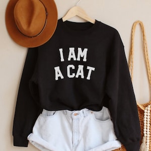 Könnte beinhalten: Schwarzes Sweatshirt mit dem Text "I AM A CAT" in weißen Buchstaben. Das Sweatshirt wird mit hellblauen Jeans-Shorts und einem braunen Hut getragen.