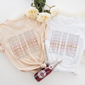 May include: Two t-shirts with the text "Let's Go Girls" repeated in a gradient of pink and orange. The shirts are laid out on a white surface with a bottle of champagne.