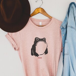 May include: A pink t-shirt with a black frog graphic and the word "MOOD" printed below it. The shirt is styled with a brown hat and a light blue denim jacket.