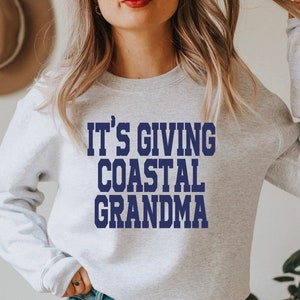 Könnte beinhalten: Ein graues Sweatshirt mit dem Text "IT'S GIVING COASTAL GRANDMA" in Blau gedruckt.