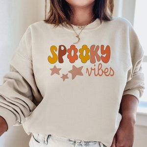May include: A beige sweatshirt with the text "Spooky Vibes" in a retro font with orange, yellow, and pink colors. The text is surrounded by three pink stars.