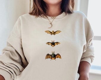 May include: A beige sweatshirt with a graphic of three bats in different sizes. The bats are brown, green, and yellow. The sweatshirt is a relaxed fit and has a crew neck.