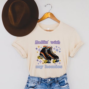 May include: A light beige t-shirt with a graphic of roller skates and the text "Rollin' with my homies". The shirt is being displayed on a hanger with a brown hat on top.