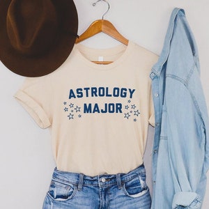 K&ouml;nnte beinhalten: Ein hellbeiges T-Shirt mit dem Text "Astrology Major" in blauer Schrift und kleinen blauen Sternen. Das Shirt h&auml;ngt an einem Holzb&uuml;gel mit einem braunen Hut und einer hellblauen Jeansjacke.