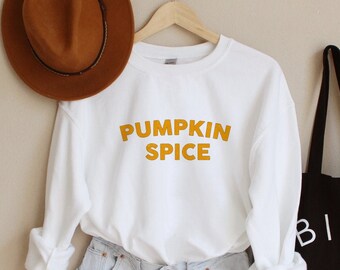 May include: White long-sleeved sweatshirt with the text "Pumpkin Spice" in orange lettering. The sweatshirt is paired with light blue denim shorts and a brown fedora. A black tote bag with the text "Bien Fait" in white lettering is also pictured.