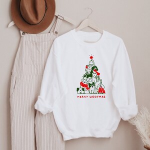 May include: White sweatshirt with a Christmas tree design made of dogs, with green and red accents. The text "MERRY WOOFMAS" is printed below the tree. A brown hat and striped overalls hang in the background.