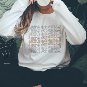 May include: White sweatshirt with the phrase "Let's Go Girls" repeated in a pink and orange retro font.