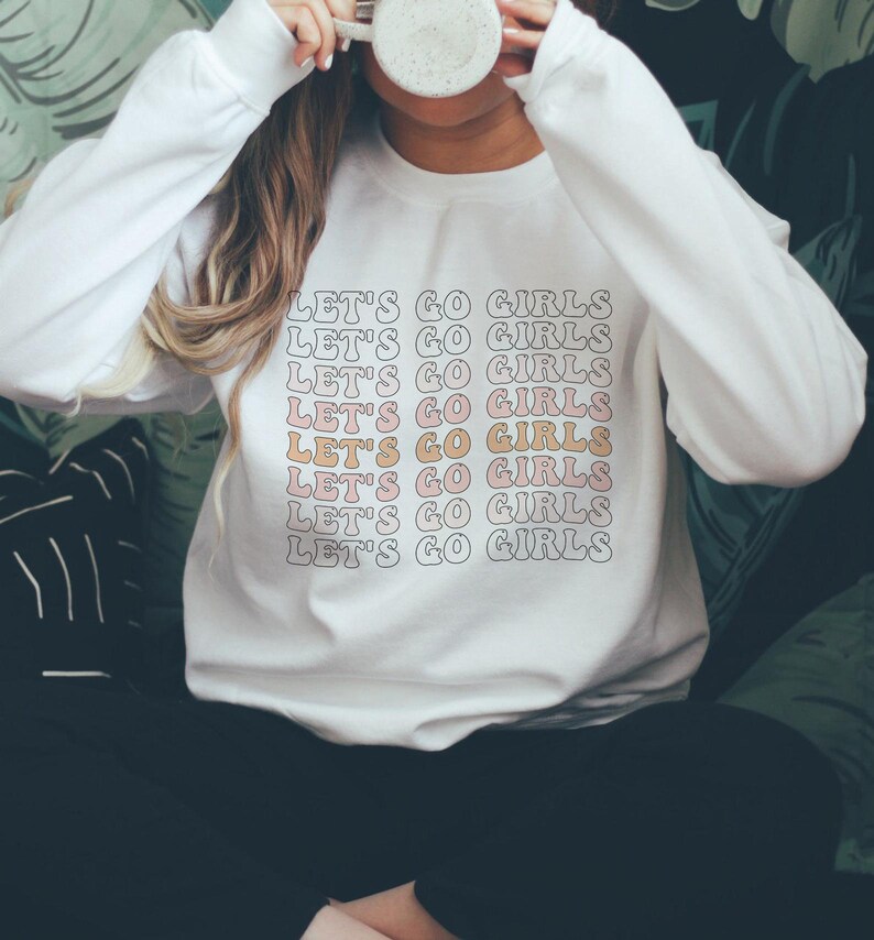May include: White sweatshirt with the phrase "Let's Go Girls" repeated in a pink and orange retro font.