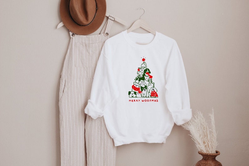 May include: White sweatshirt with a Christmas tree design made of dogs, with green and red accents. The text "MERRY WOOFMAS" is printed below the tree. A brown hat and striped overalls hang in the background.