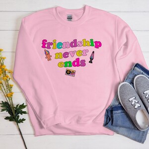 May include: Pink sweatshirt with the text "friendship never ends" in rainbow colors. The text is surrounded by a troll doll, a lava lamp, and a cassette tape.