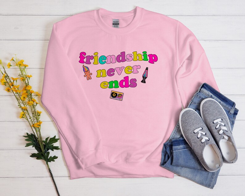 May include: Pink sweatshirt with the text "friendship never ends" in rainbow colors. The text is surrounded by a troll doll, a lava lamp, and a cassette tape.