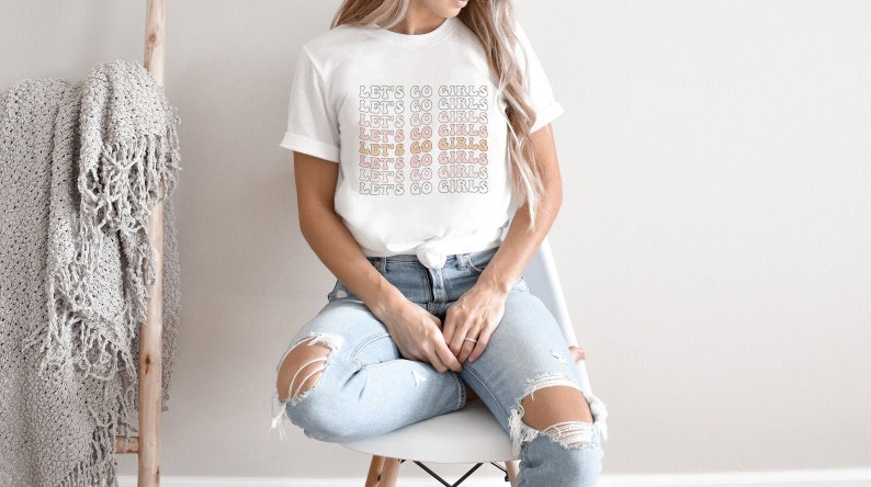 May include: A white t-shirt with the text "Let's Go Girls" repeated in a gradient of pink, orange, and yellow.
