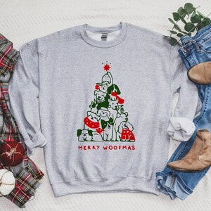 May include: A light gray sweatshirt featuring a Christmas tree design made of various dog breeds. The tree is topped with a red star and the words "MERRY WOOFMAS" are printed in red. The sweatshirt is paired with blue jeans and brown boots.
