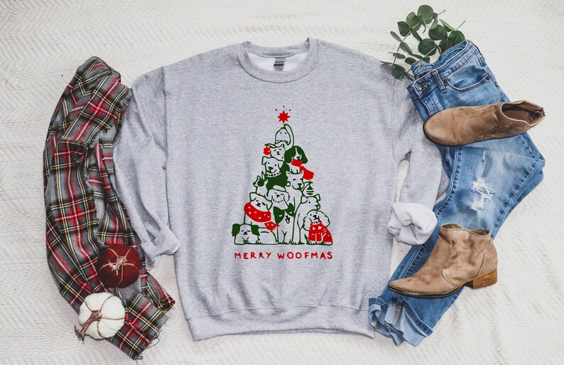 May include: A light gray sweatshirt featuring a Christmas tree design made of various dog breeds. The tree is topped with a red star and the words "MERRY WOOFMAS" are printed in red. The sweatshirt is paired with blue jeans and brown boots.