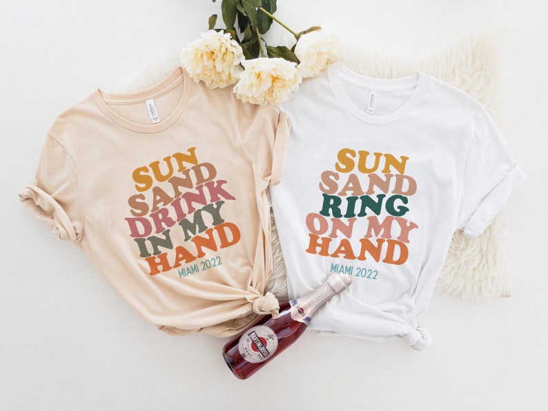 May include: Two white t-shirts with a colorful graphic design that says "SUN SAND DRINK IN MY HAND MIAMI 2022" and "SUN SAND RING ON MY HAND MIAMI 2022".