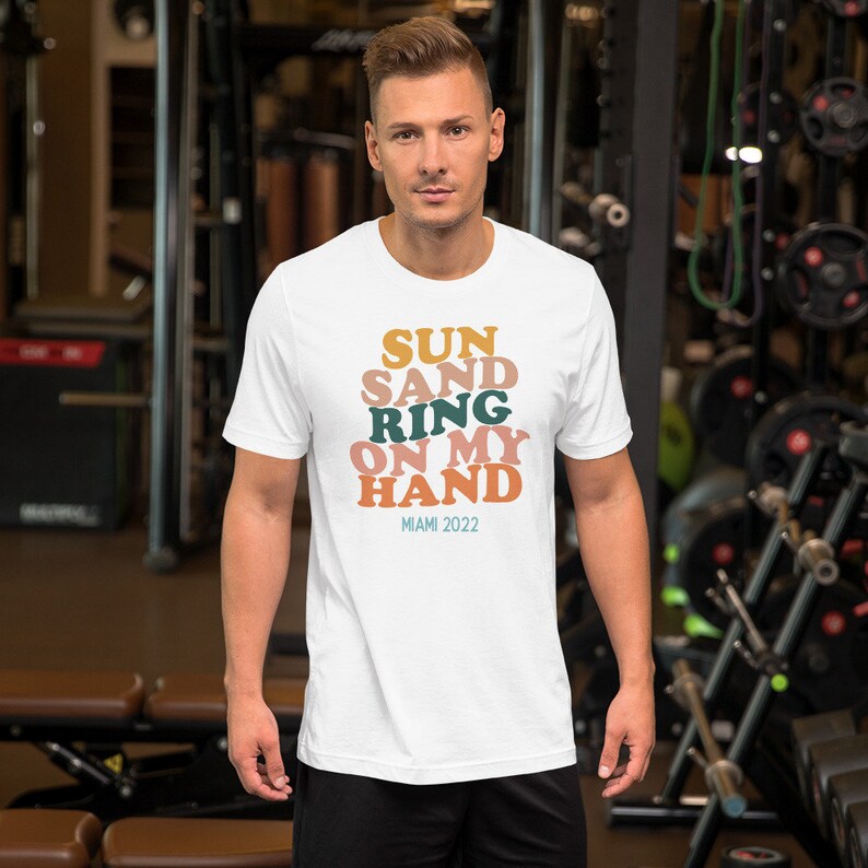 May include: White t-shirt with a graphic design that says "SUN SAND RING ON MY HAND" in colorful, stacked letters. The text "MIAMI 2022" is printed below the graphic.