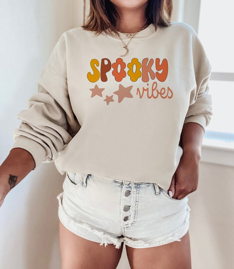 May include: A beige sweatshirt with the text "Spooky Vibes" in a retro font with orange, yellow, and pink colors. The text is surrounded by three pink stars.