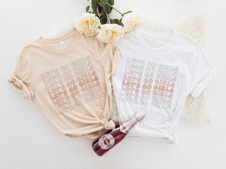May include: Two t-shirts with the text "Let's Go Girls" repeated in a gradient of pink and orange. The shirts are laid out on a white surface with a bottle of champagne.